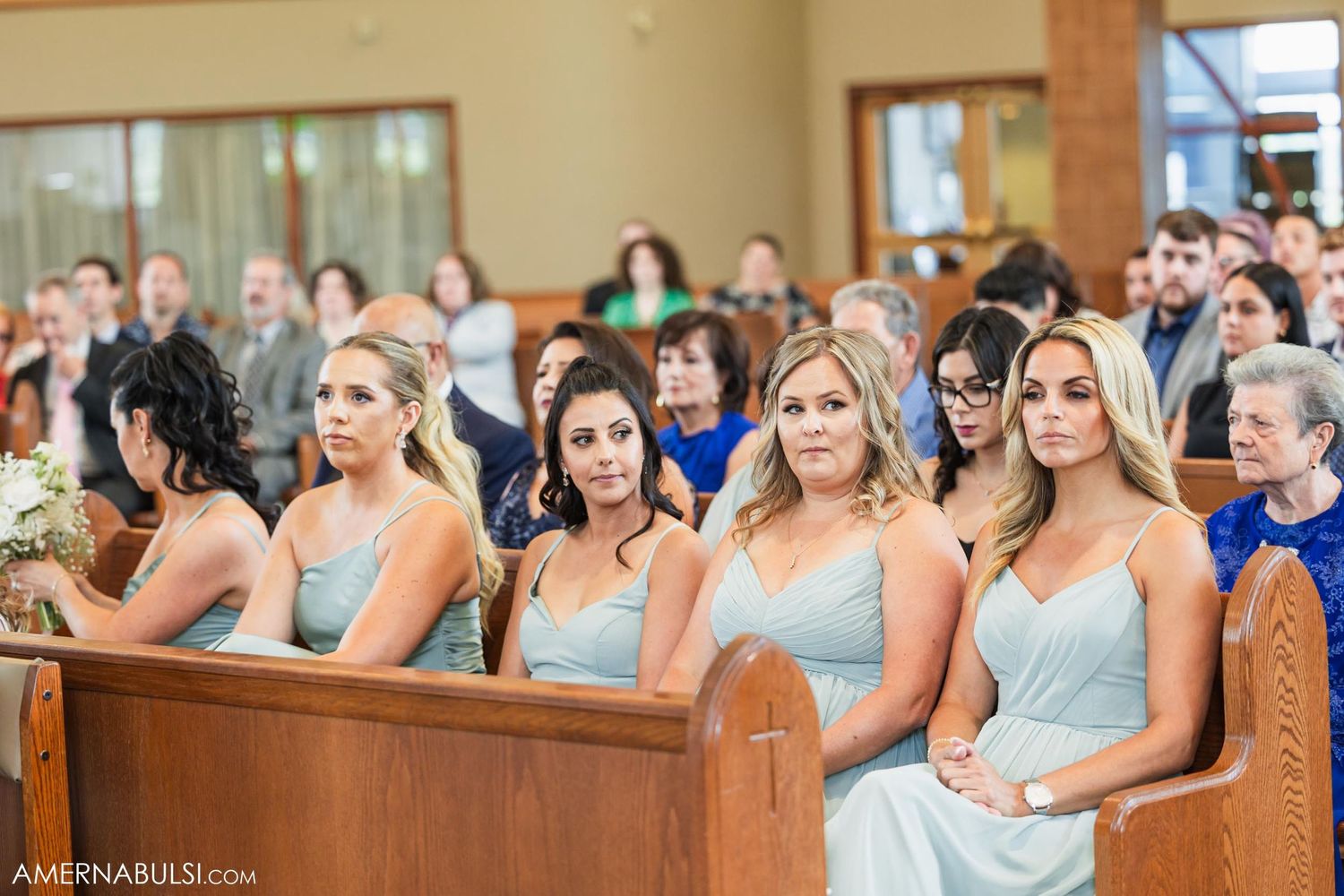 Bridesmaids in light blue dresses sit in wooden church pews during a wedding ceremony.