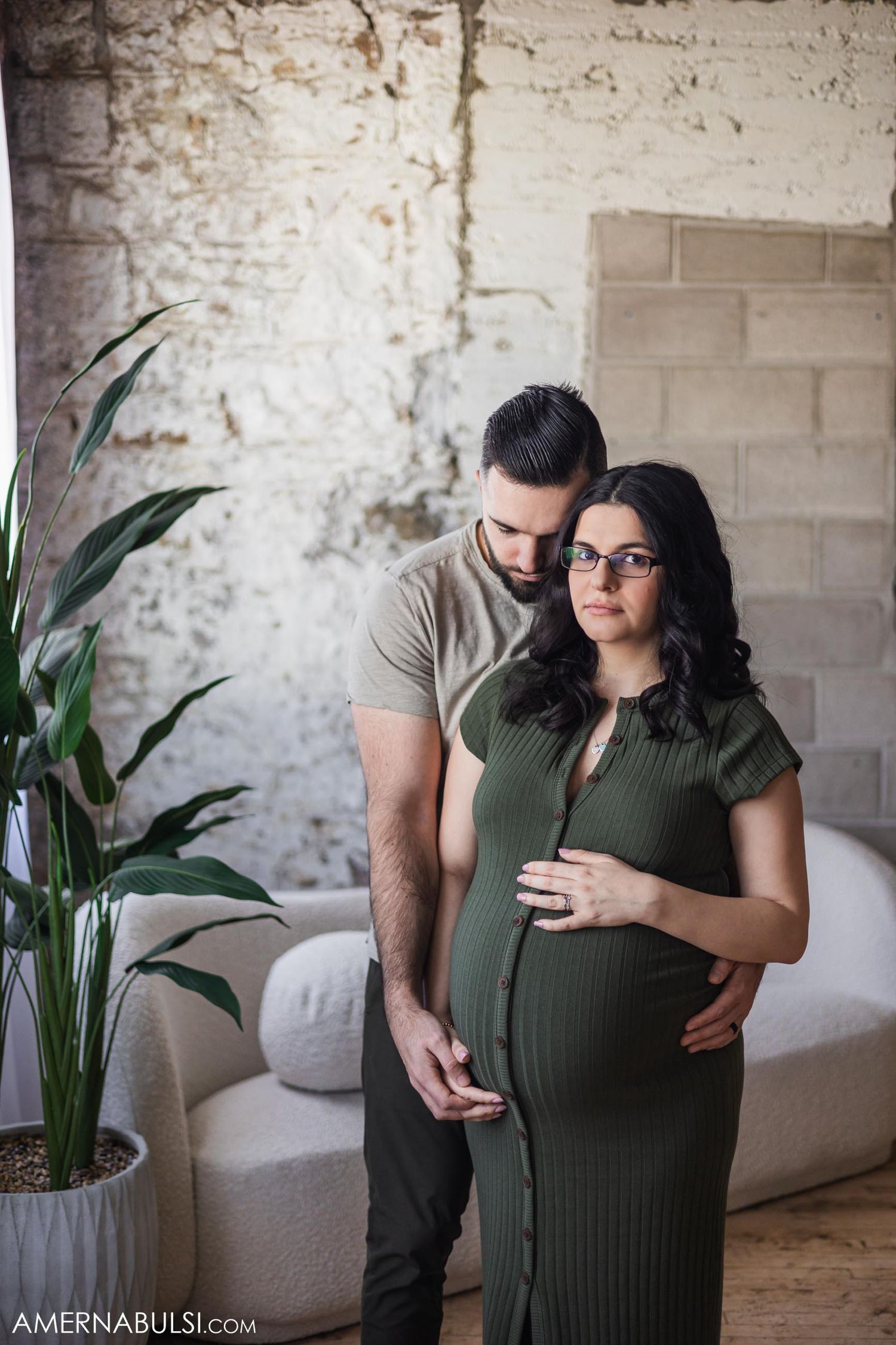 Intimate maternity photoshoot in olive dress against rustic white brick wall with indoor plant.