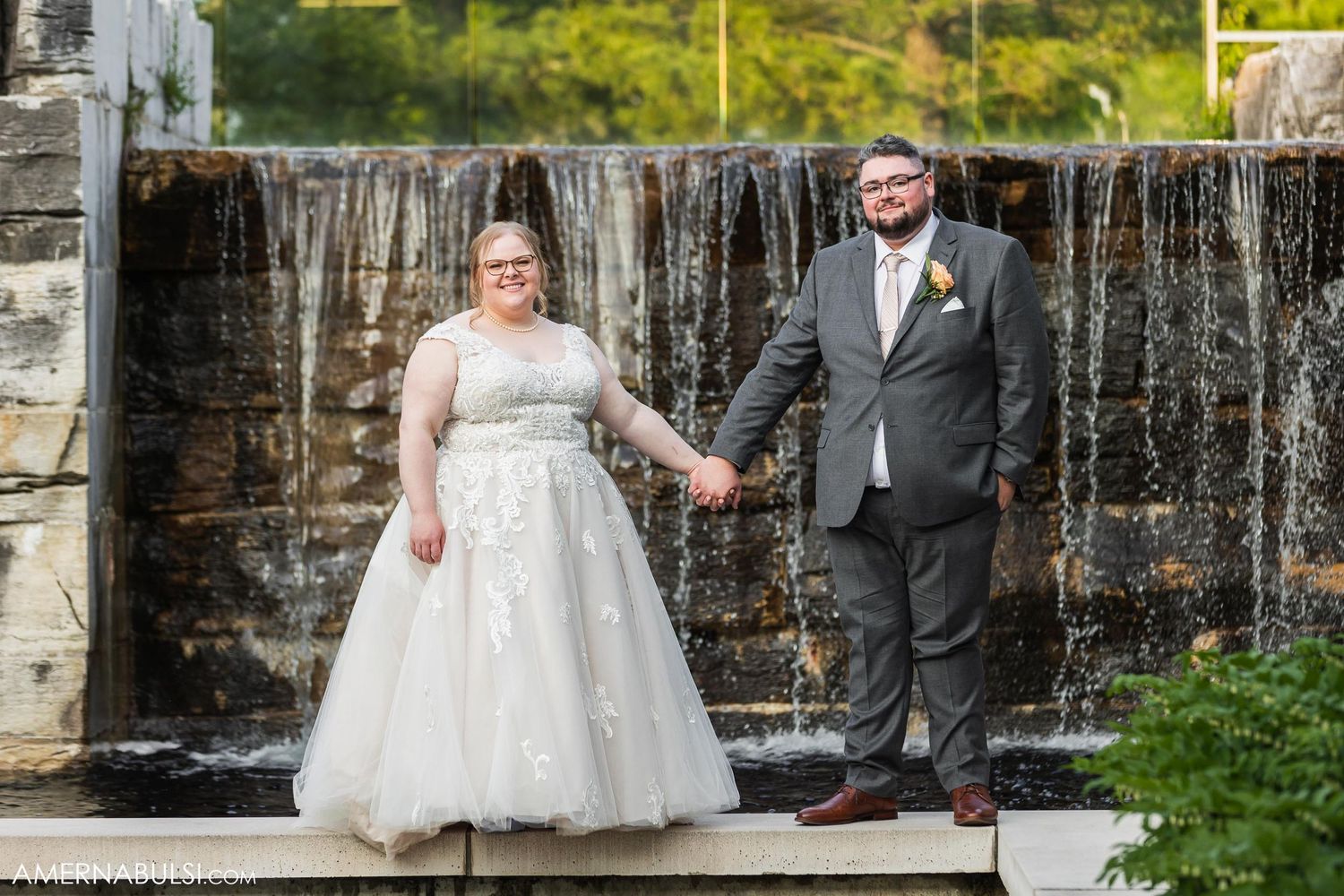 A couple poses in wedding attire in front of a cascading waterfall backdrop.