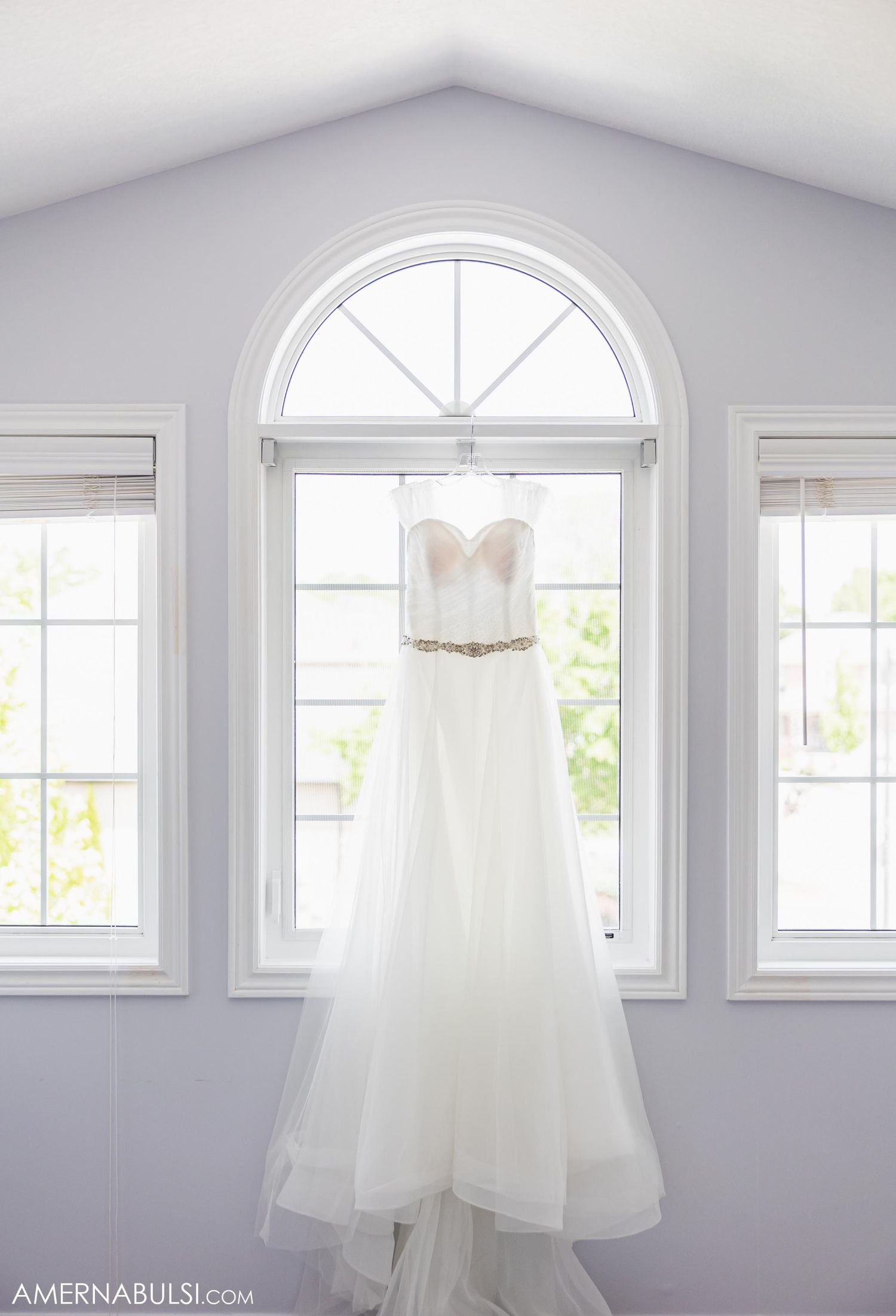 A strapless white wedding gown with beaded belt hangs elegantly in front of arched window.