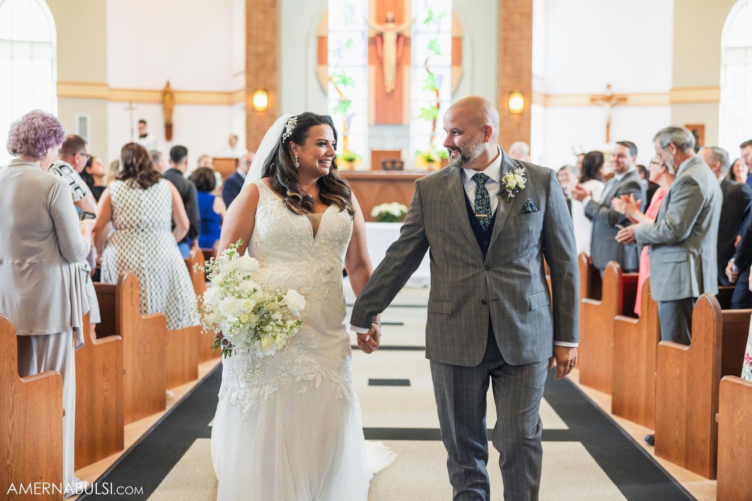 Couple walks down church aisle holding white floral bouquet.