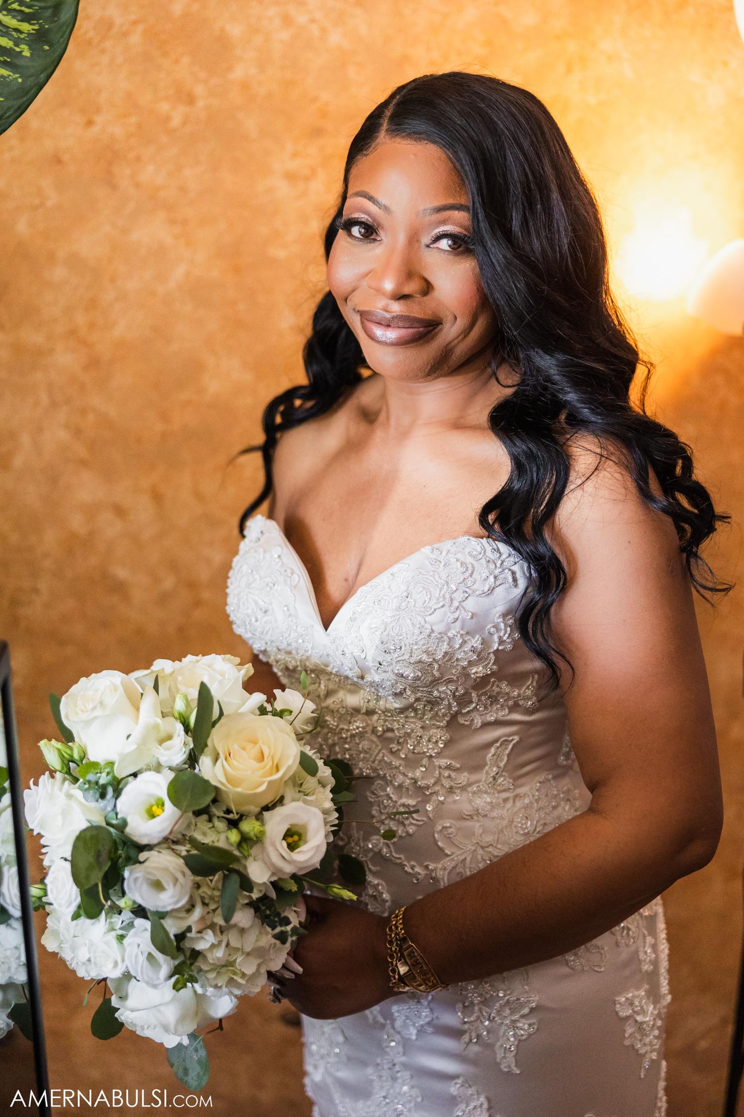 Bride in lace wedding dress holds white rose bouquet against golden wall.