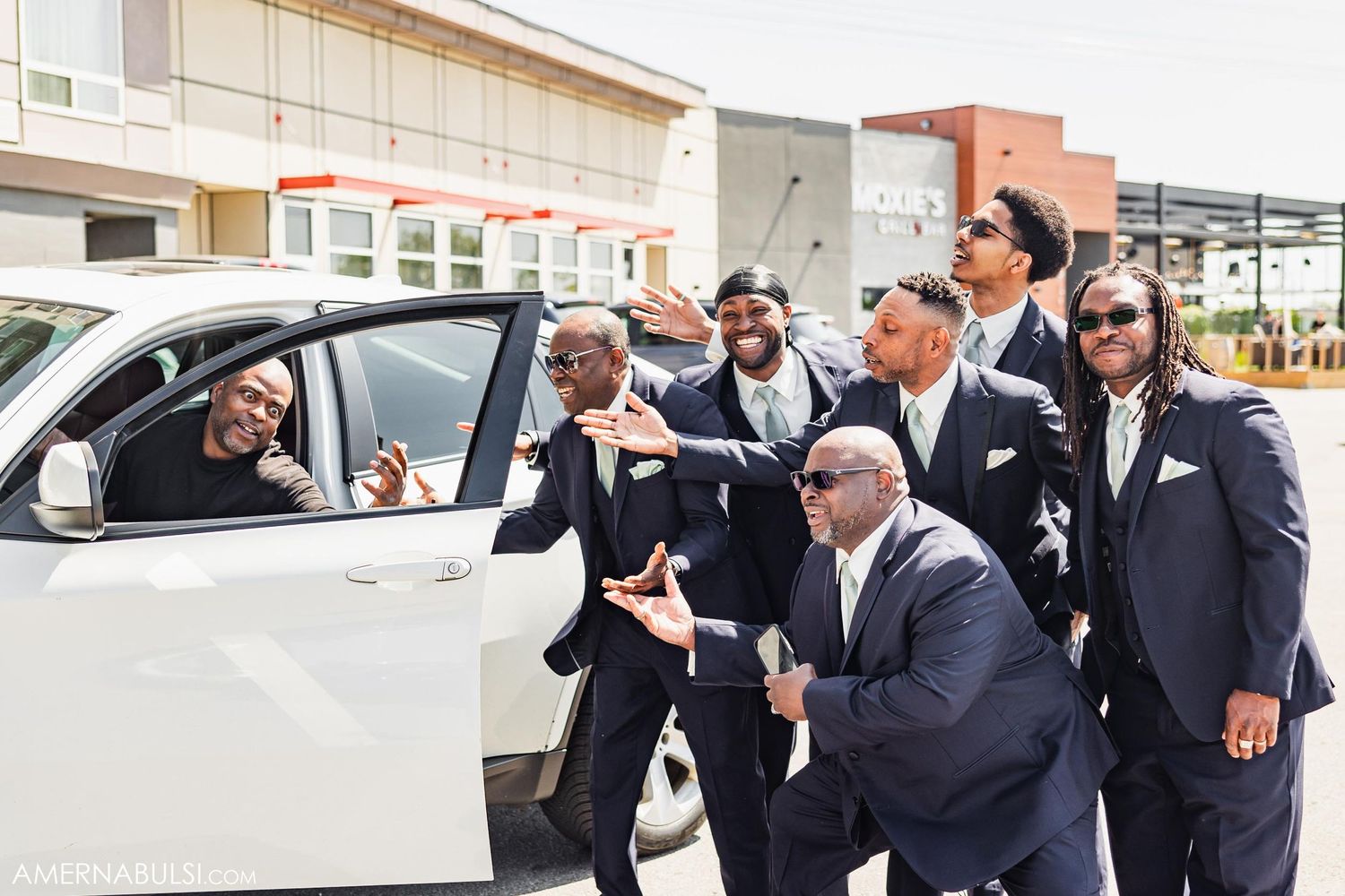 Groomsmen in suits gather around a white car, sharing a joyful moment before the wedding.