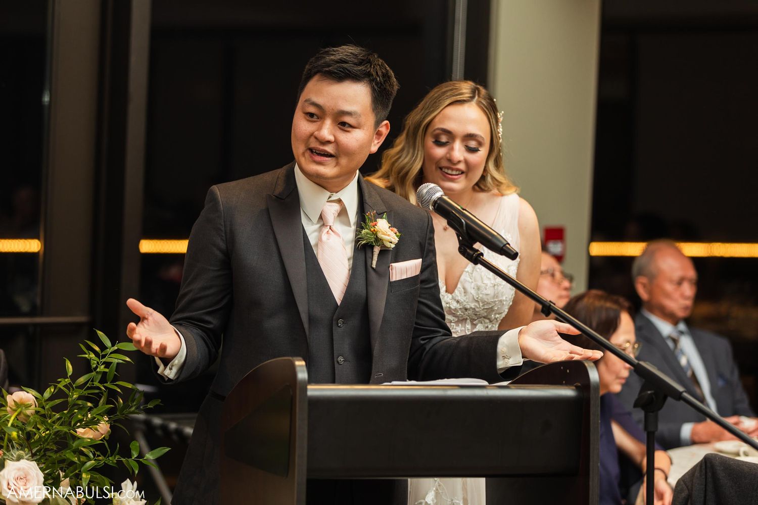 Wedding reception speech being delivered at a decorated head table with floral arrangements.