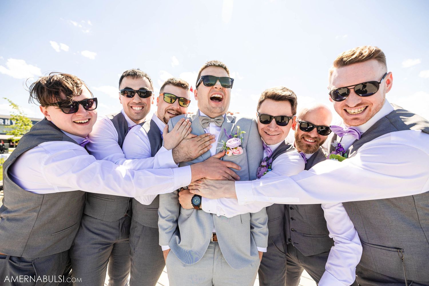 Playful groomsmen in grey suits and sunglasses huddling together outdoors.