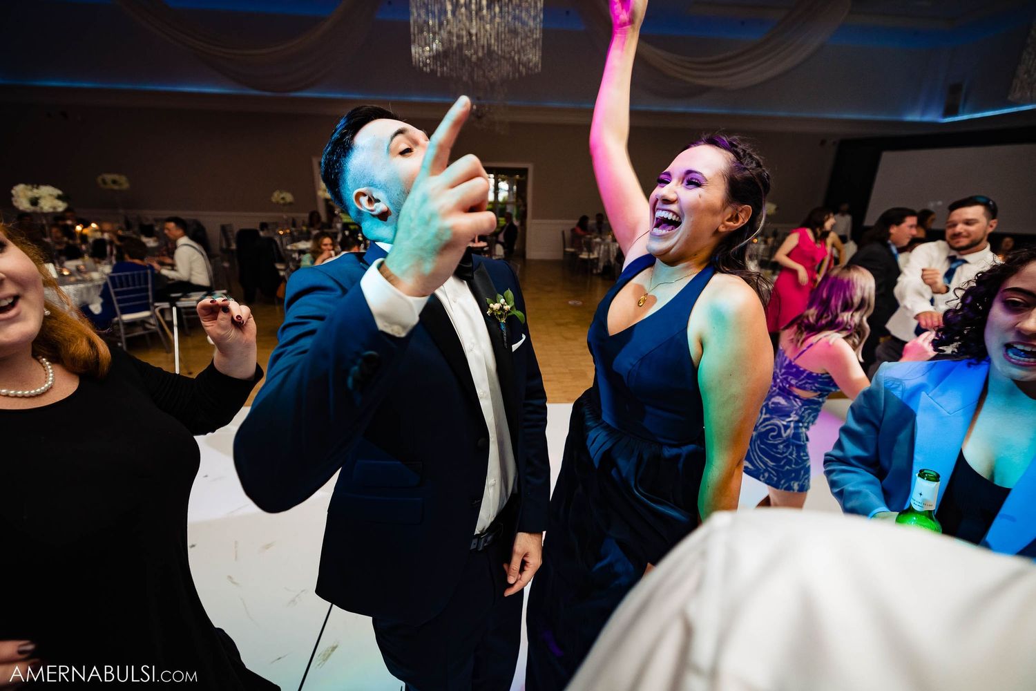 Wedding guests dance enthusiastically on a crowded reception dance floor under colorful lights and chandeliers.
