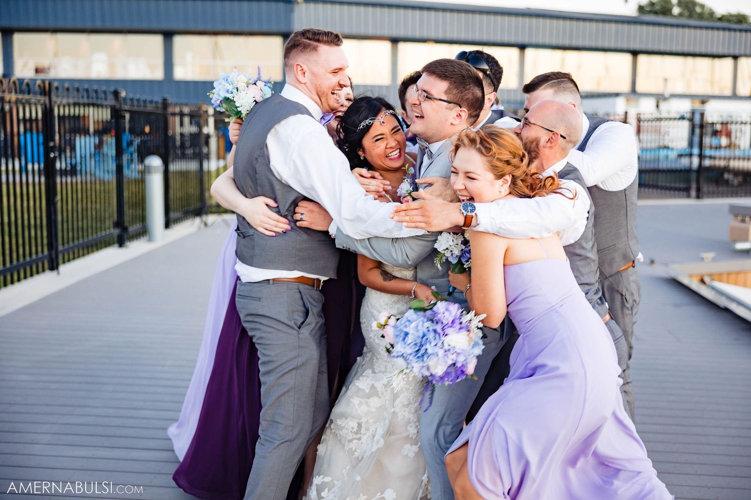 Wedding group embracing in playful pose on waterfront deck.