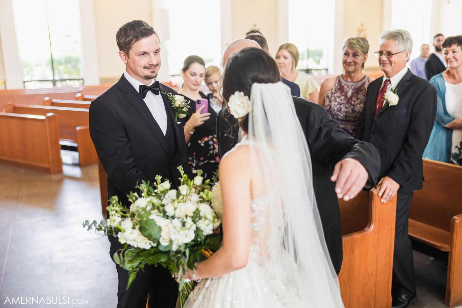 A couple exchanges vows in a church surrounded by wooden pews and floral decorations.
