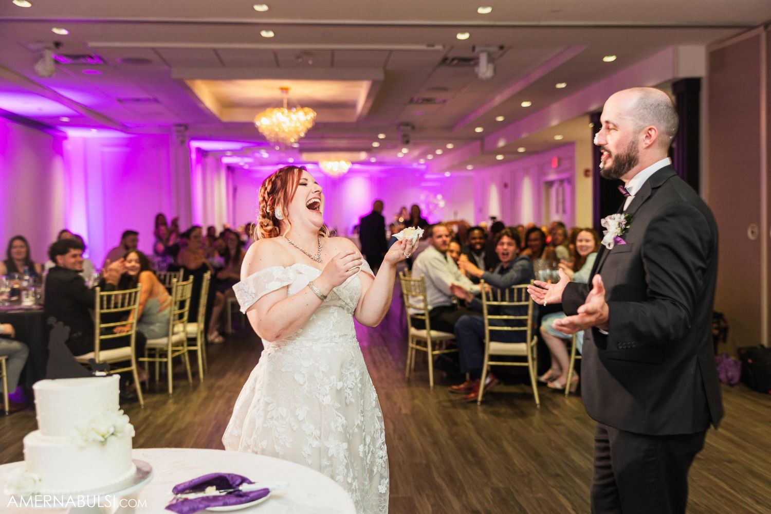 Couple sharing a playful moment while cutting their white wedding cake in purple-lit reception venue.