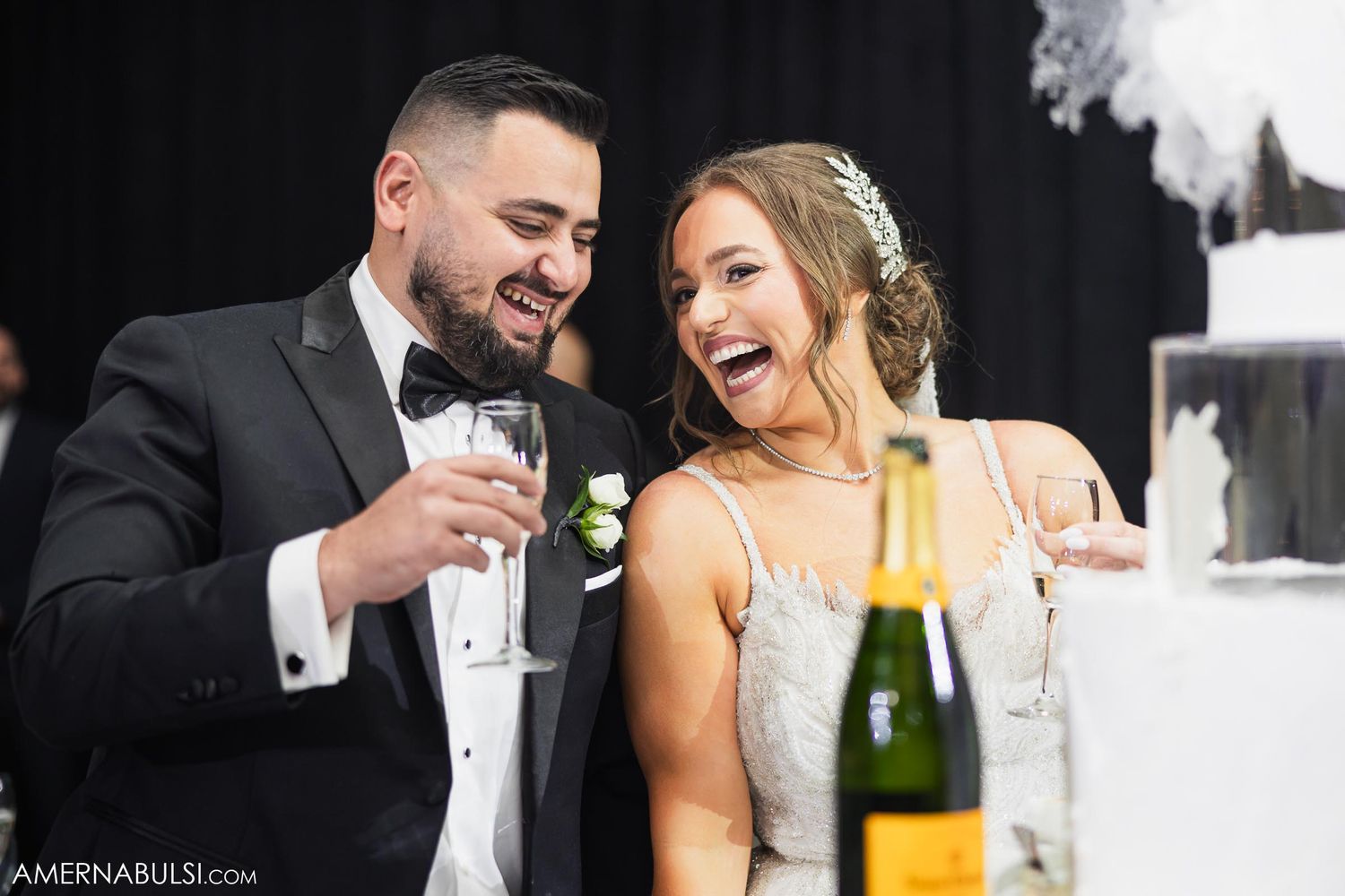 Couple shares champagne and laughter at wedding reception table.