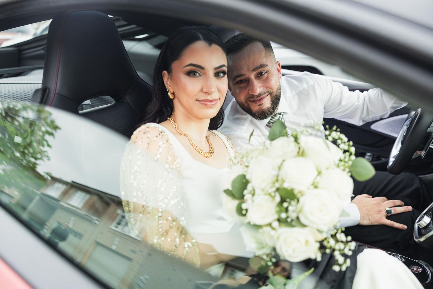 Newlyweds sitting in car with white bouquet and formal attire.