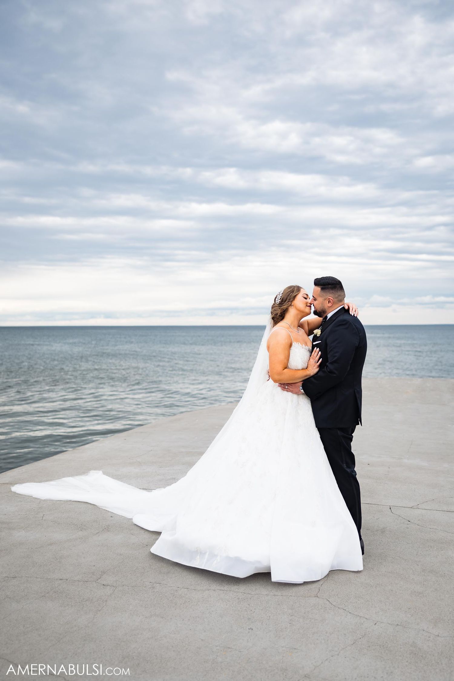 A romantic wedding couple embraces on a pier overlooking a serene ocean with dramatic cloudy skies.