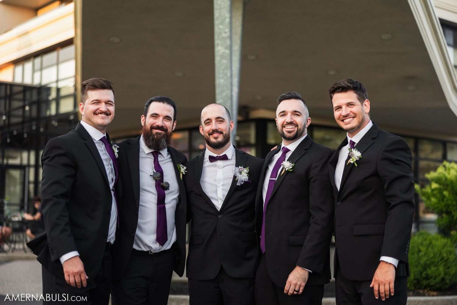 Groomsmen in black tuxedos and bowties posing together outside venue.
