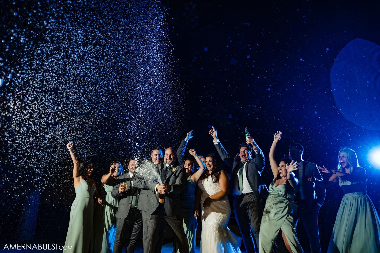 Wedding party raises their arms in celebration as confetti falls under blue stage lighting.