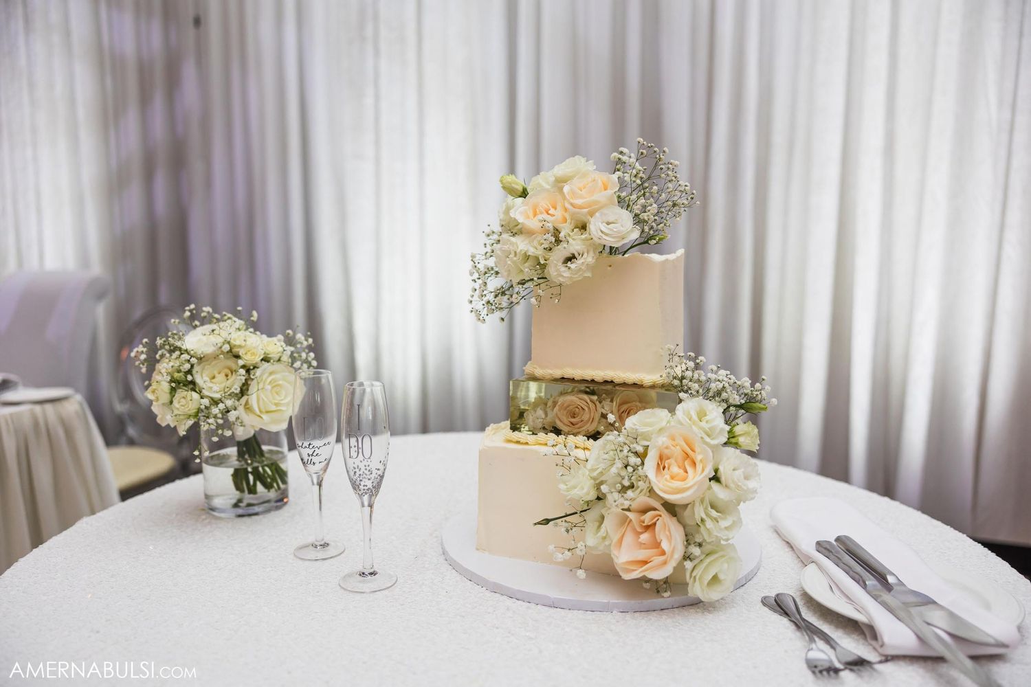 Three-tiered white wedding cake decorated with peach roses and greenery on a classic table setting.