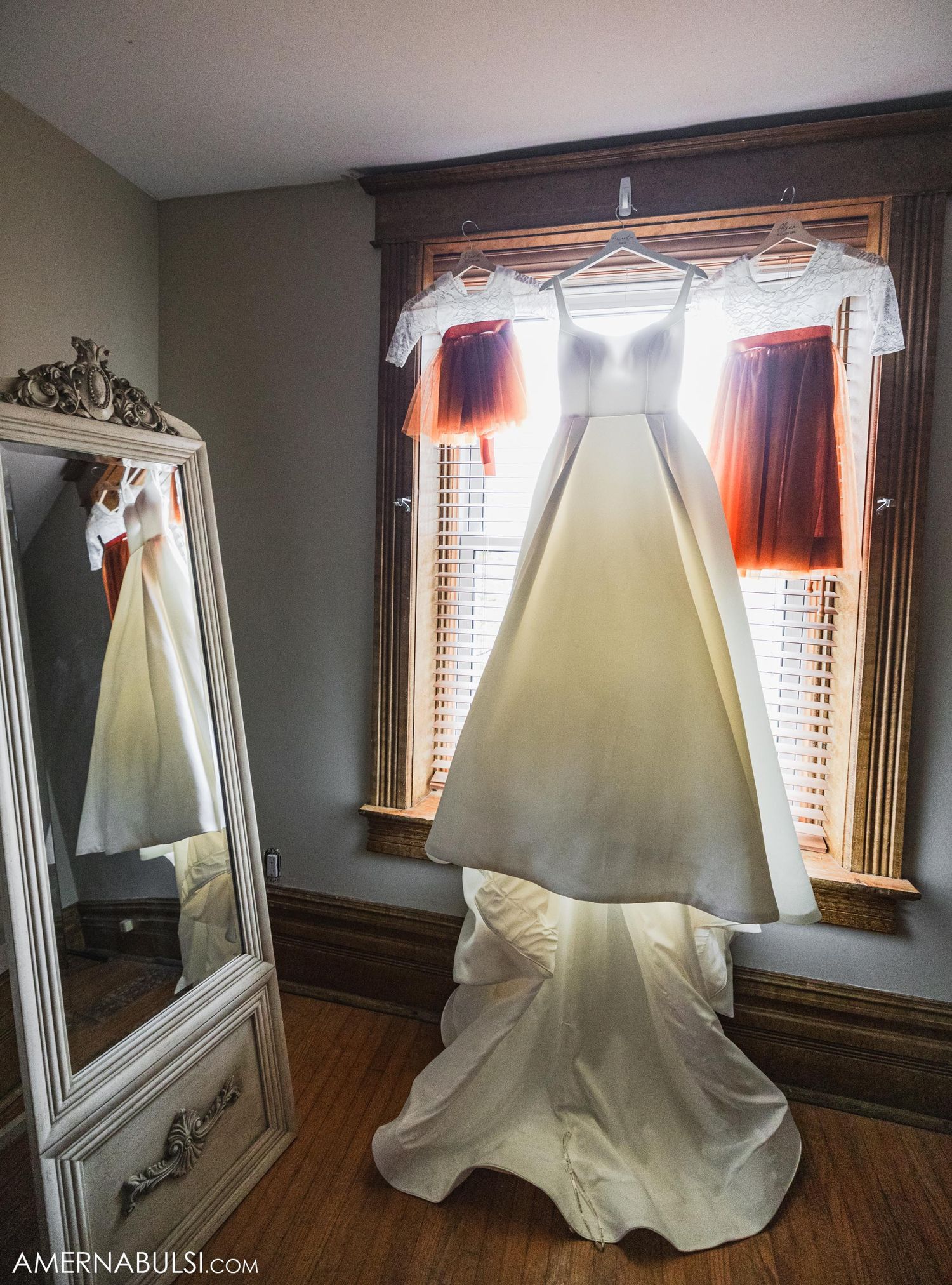 An elegant white wedding dress hanging by a window with matching bridesmaid dresses in navy and white.