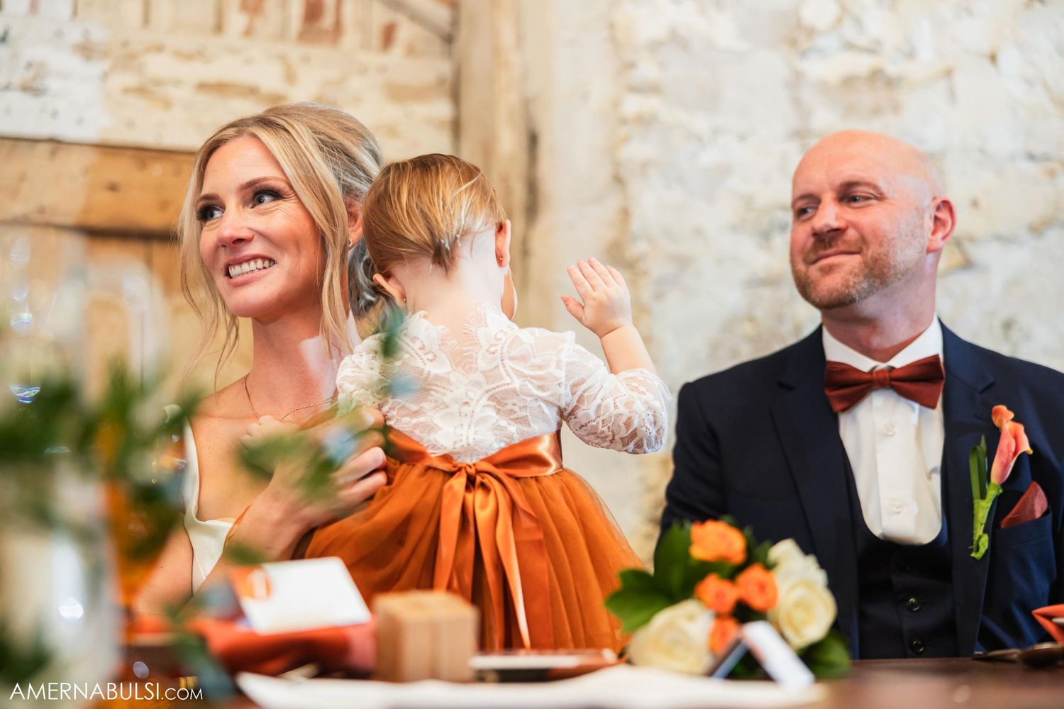 Family celebrates at rustic wedding reception with small child in orange dress at head table with floral decor.