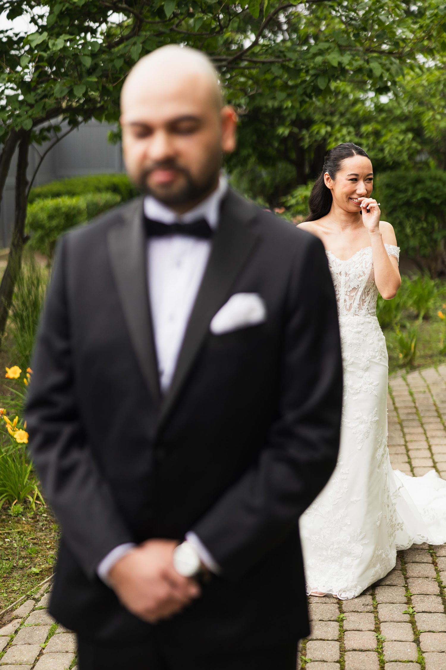 A first look wedding photo with the groom in foreground and bride in background in a garden setting.