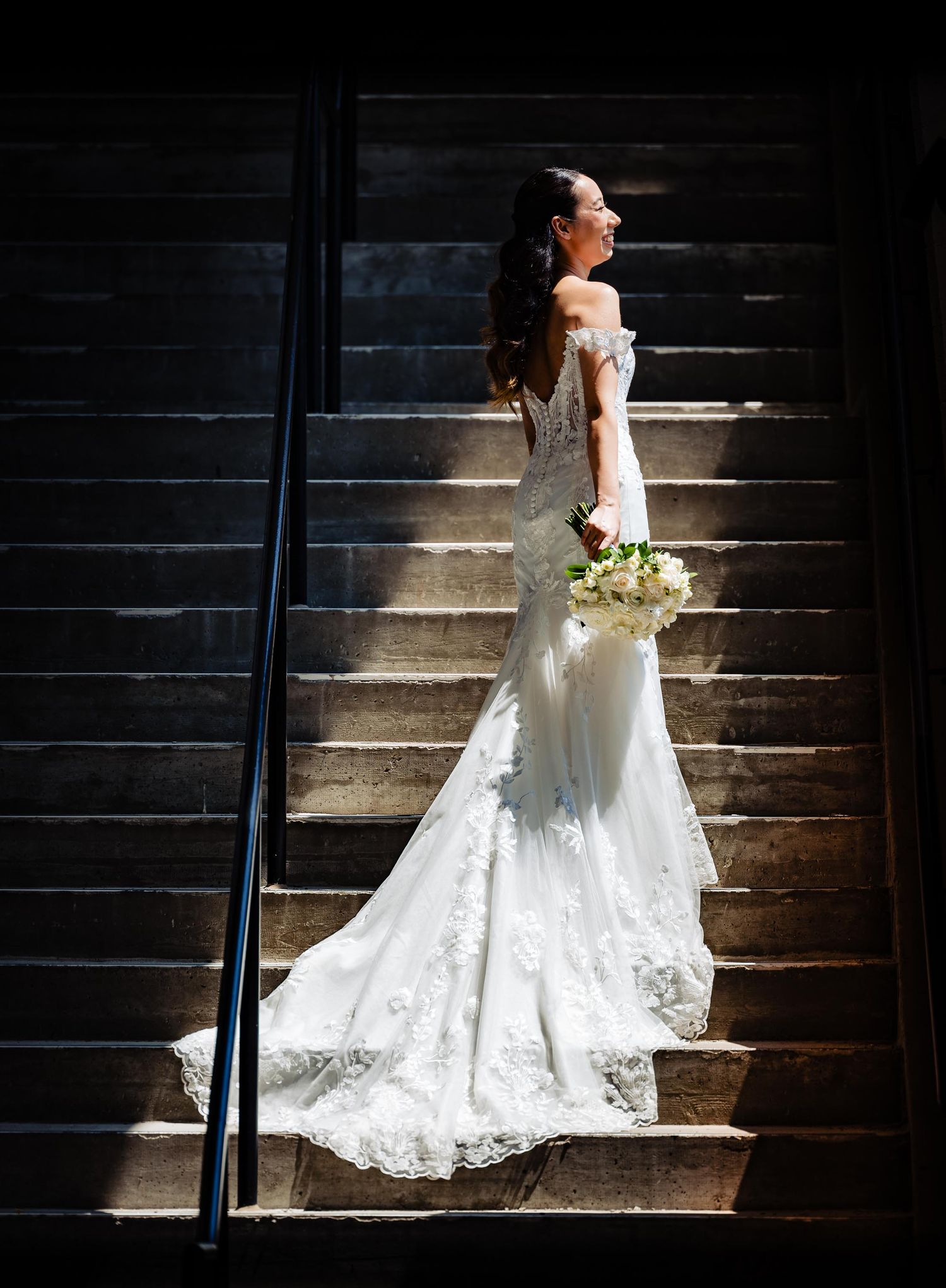 Dramatic backlit bride poses on dark stairs with flowing lace wedding dress train.
