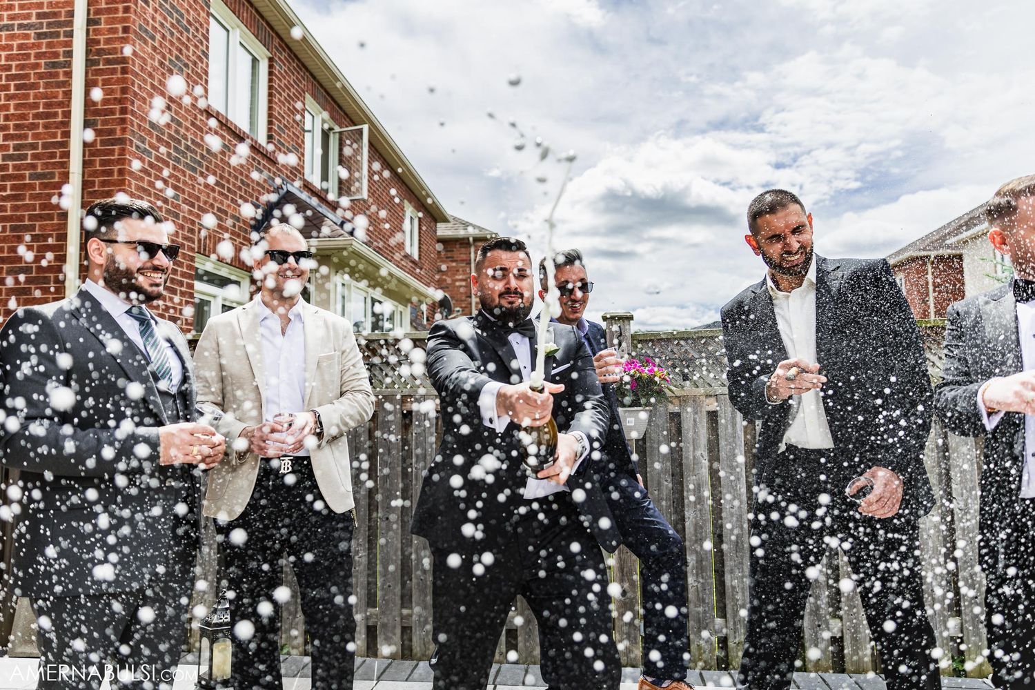 Groomsmen celebrating with champagne sprays outside a brick building.