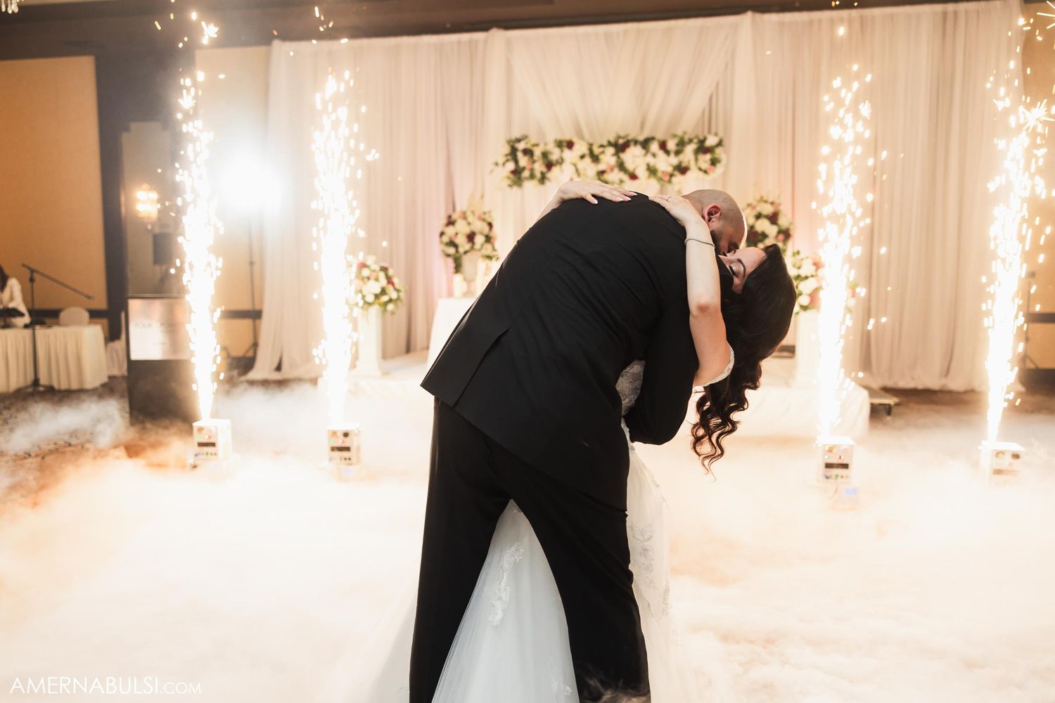 A couple shares a romantic dance moment on their wedding day surrounded by sparklers and white decor.