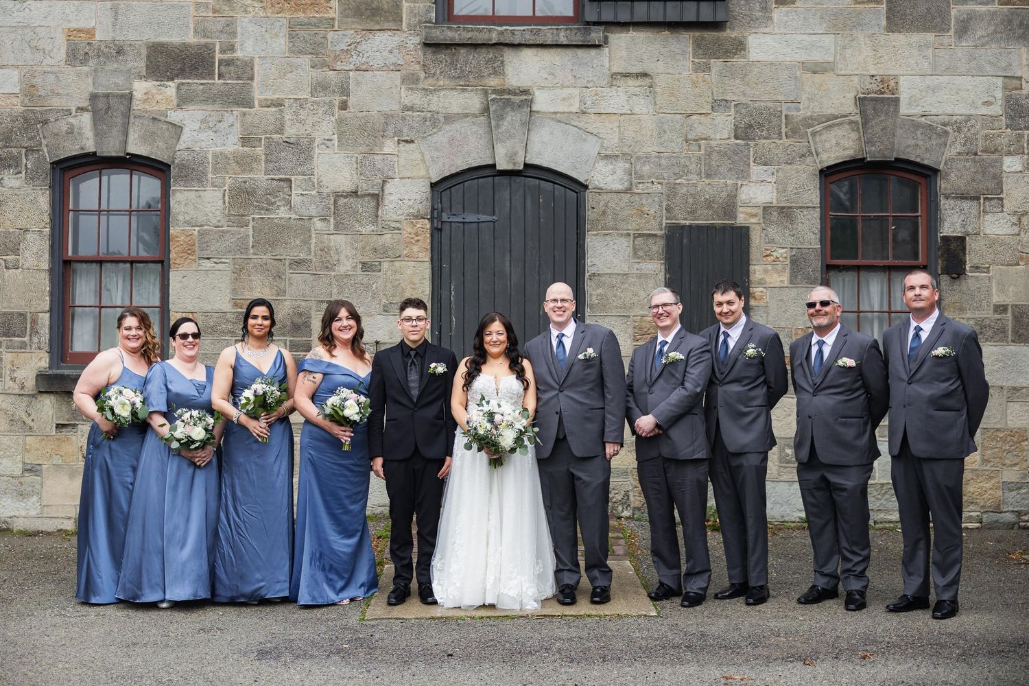 Wedding party in blue dresses and gray suits poses against stone wall backdrop.