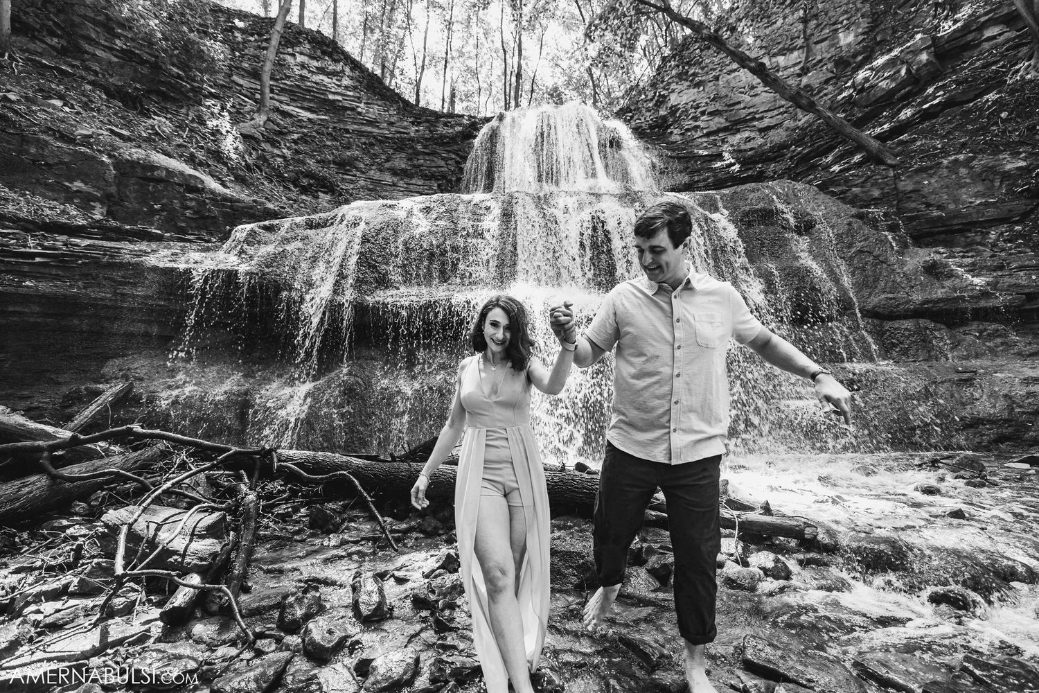 Black and white photo of two people dancing joyfully in front of a scenic waterfall.