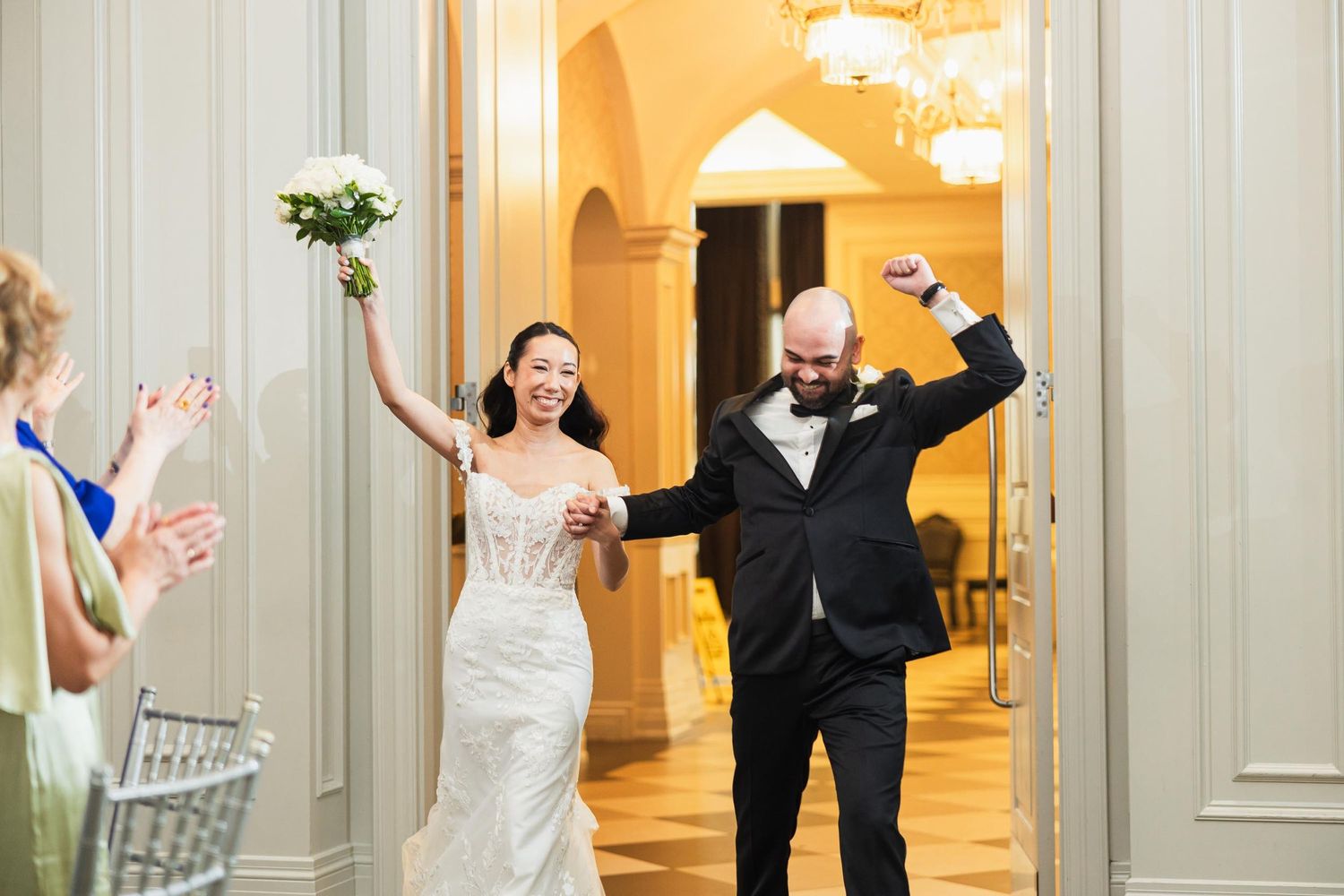 Wedding couple triumphantly enters reception hall with raised arms as guests applaud.
