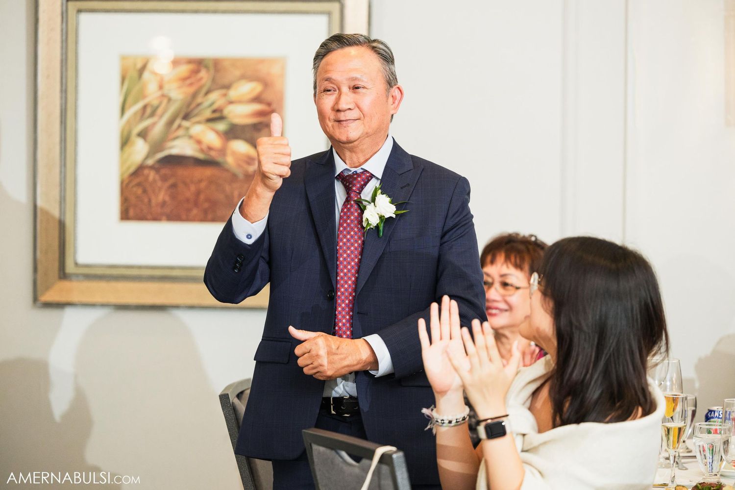 Man in navy suit delivers emotional toast during wedding reception as guests smile.