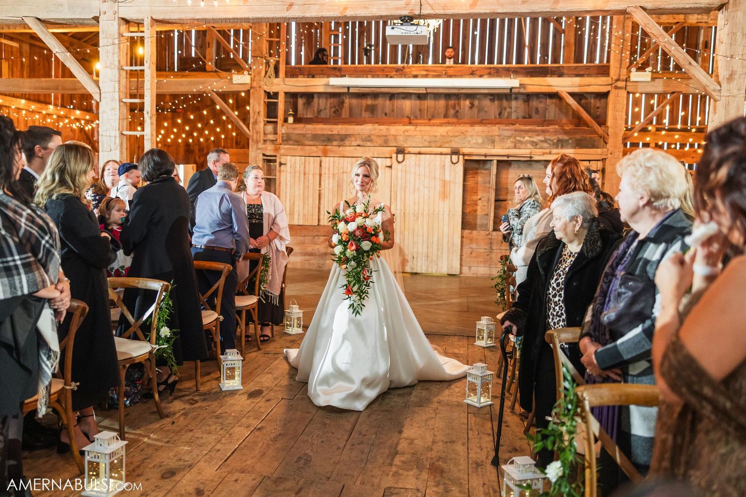 Wedding ceremony processional in rustic barn with wooden floors and string lights overhead.