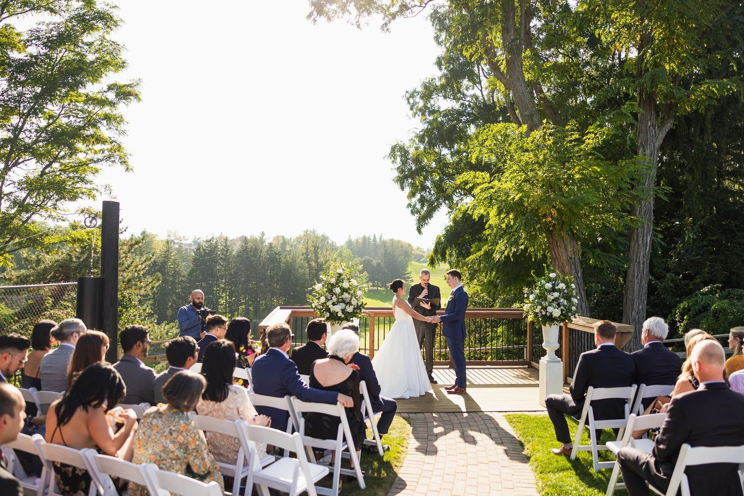 Wedding ceremony in progress under trees with guests seated in white chairs at sunset.