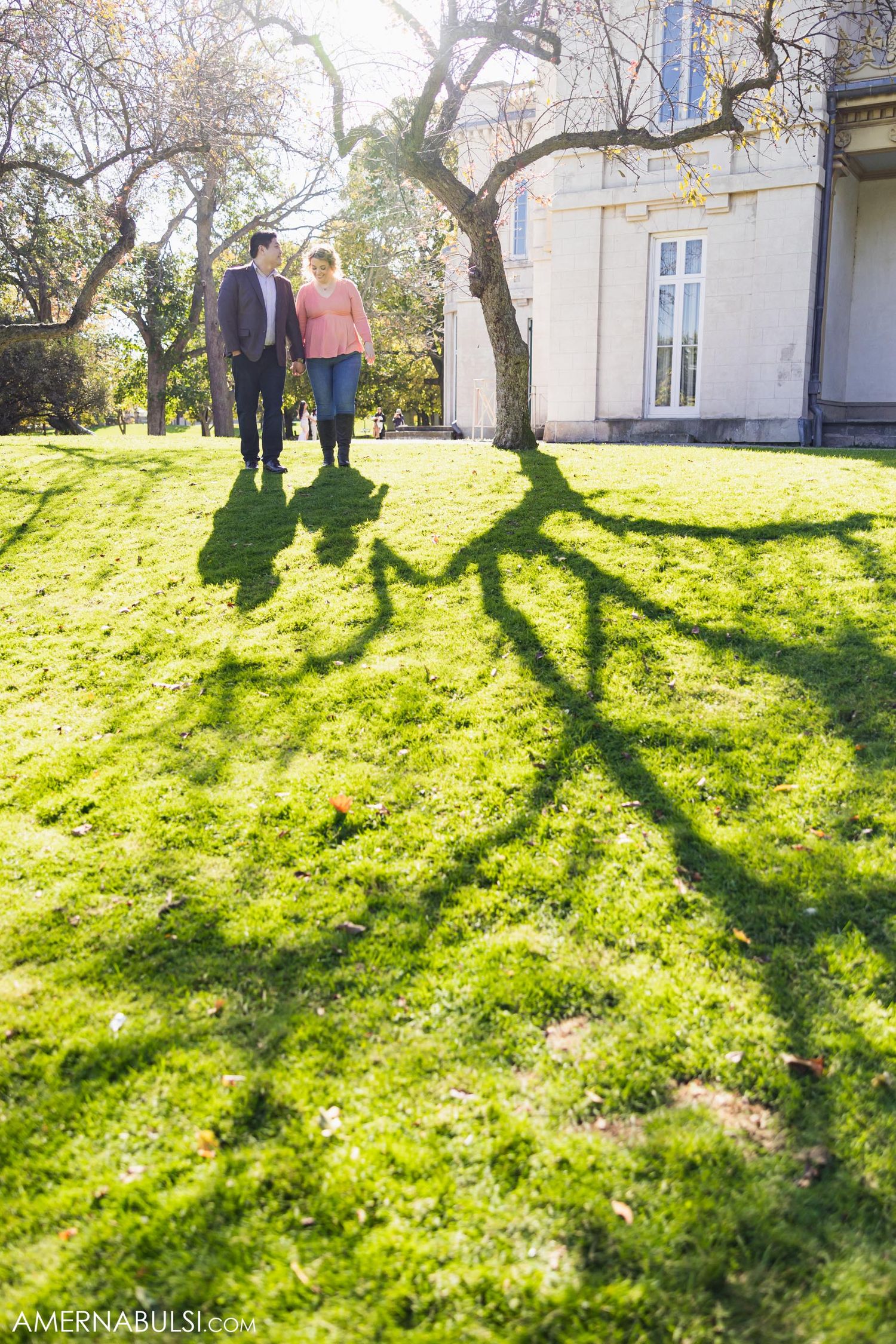 A couple walks on a sunlit lawn casting long shadows beside a historic building on a bright autumn day.