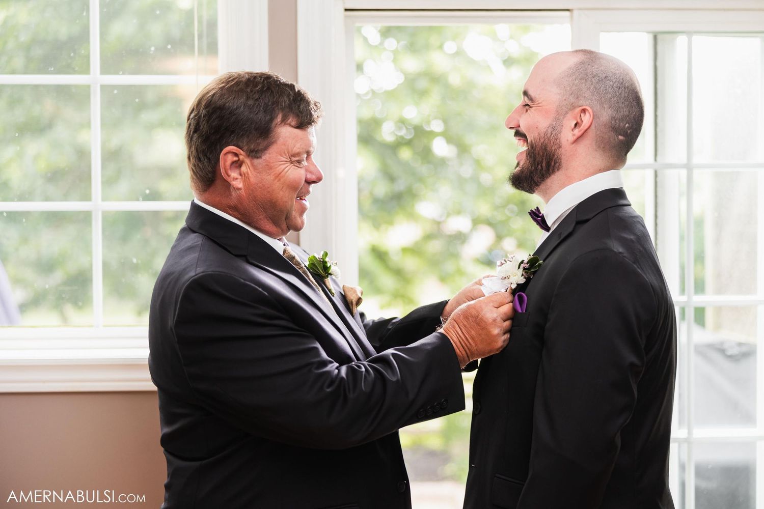Two people in black tuxedos assist with formal attire adjustments by a bright window.