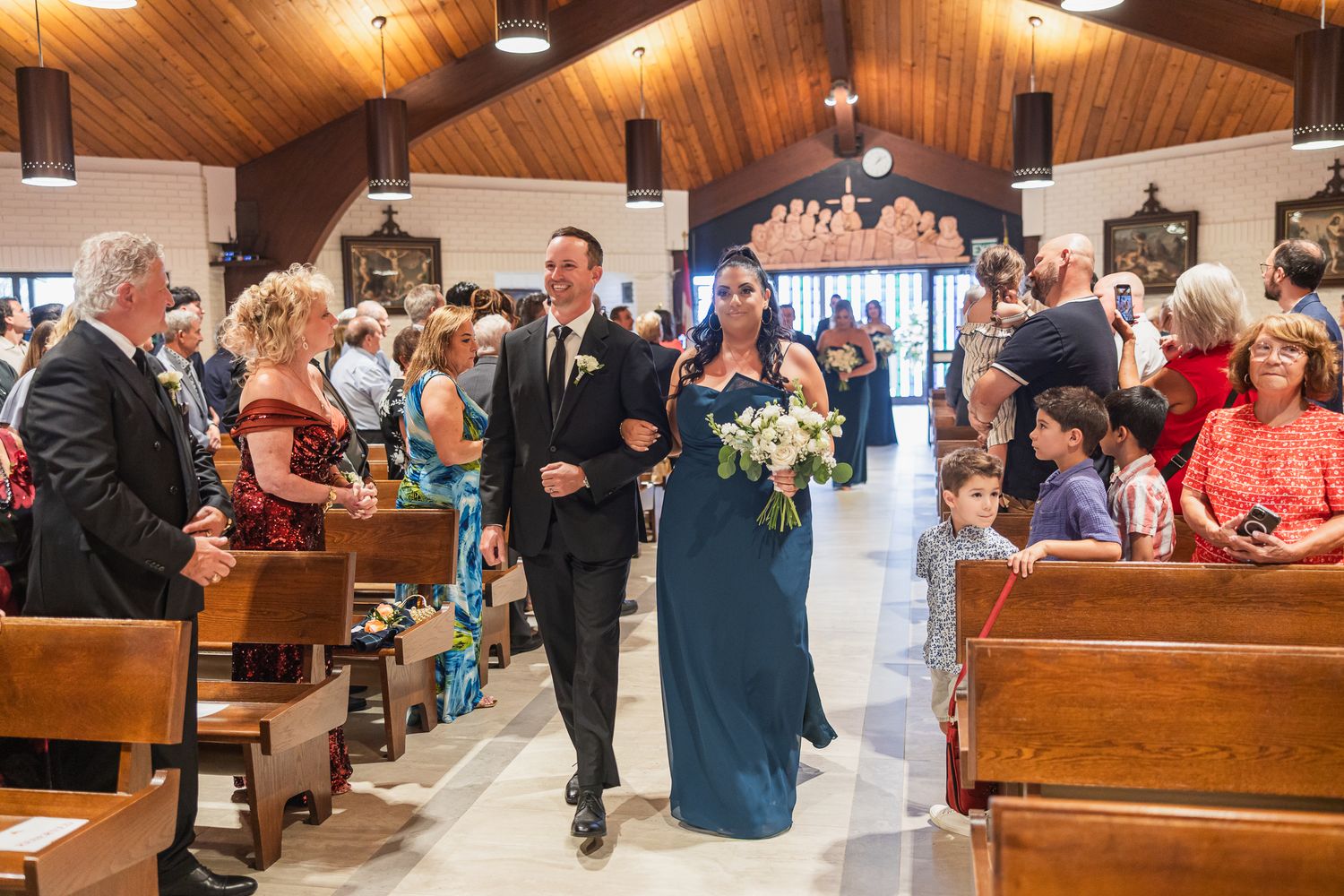 Bridesmaid in navy dress walks down the church aisle with groomsman during wedding processional.