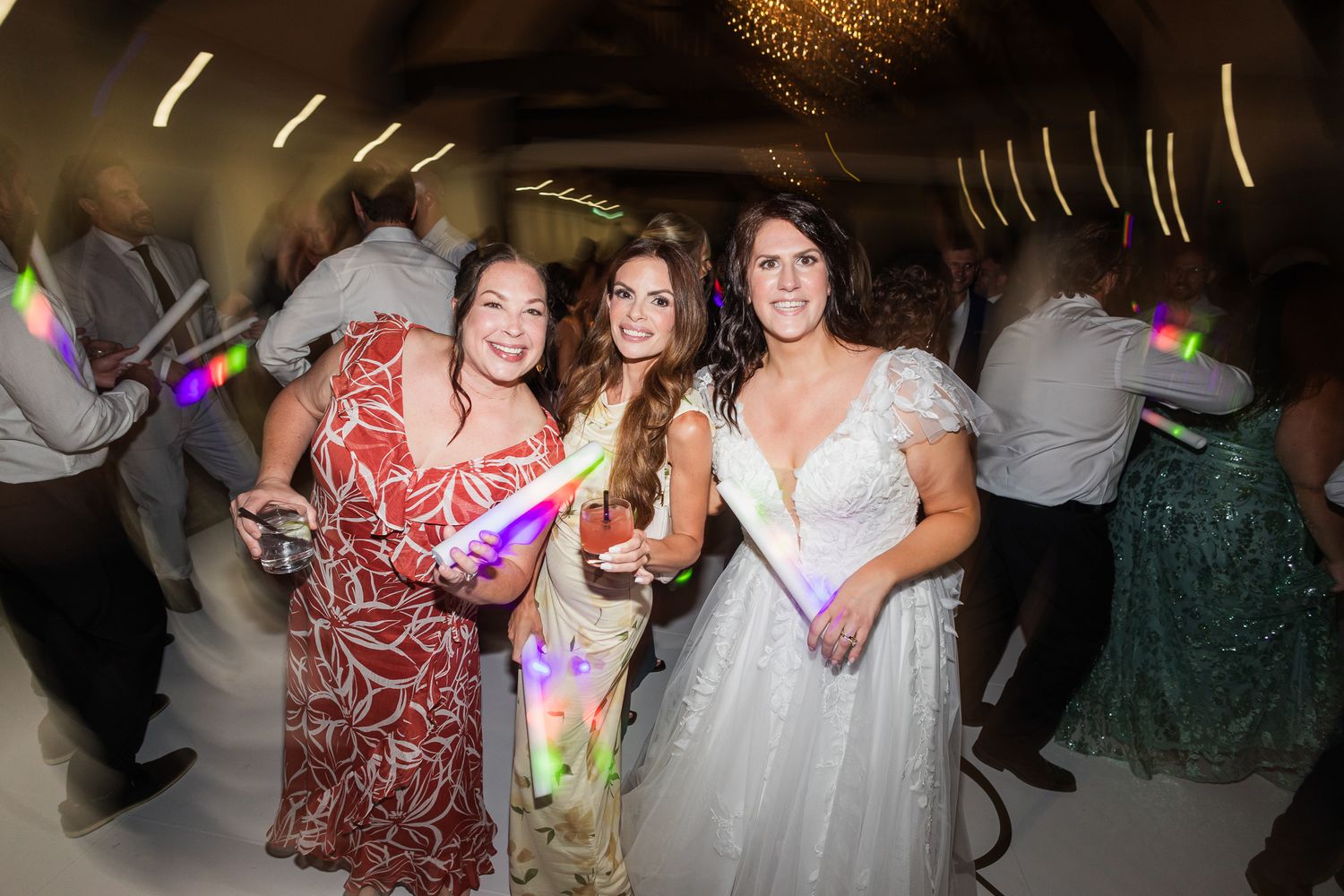 Three women smiling and posing with glow sticks at a festive wedding reception dance floor celebration.