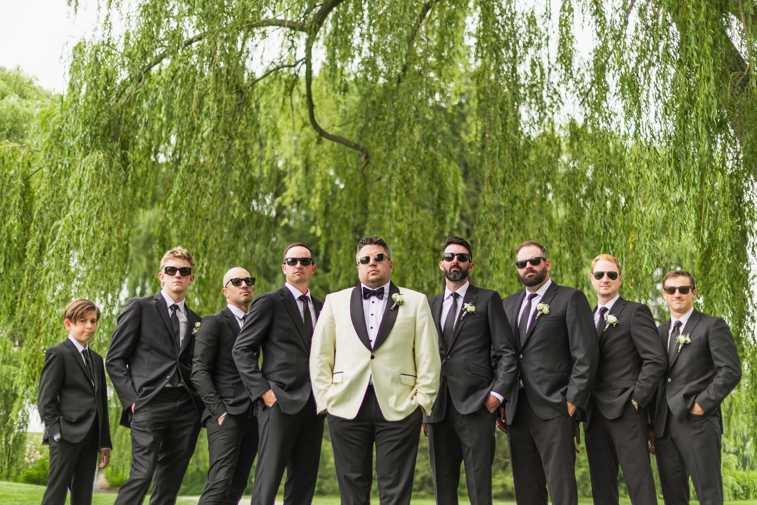 Groom in ivory tuxedo stands with groomsmen in black suits and sunglasses under a weeping willow tree.