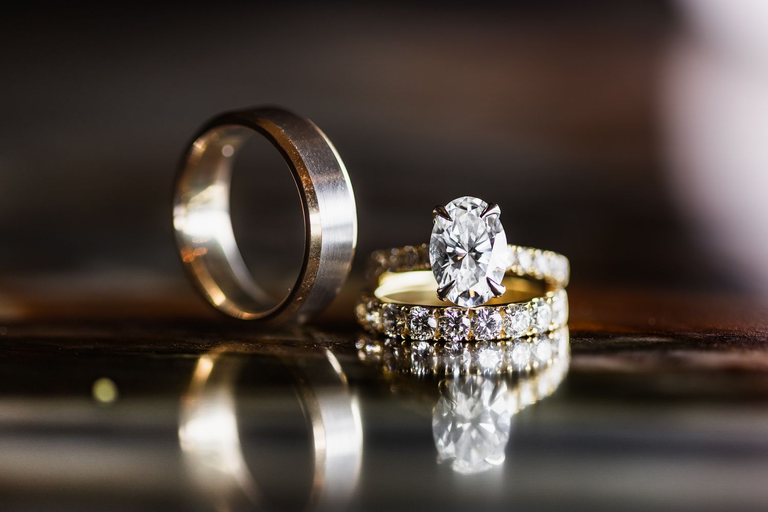 Gold oval diamond engagement ring paired with a wedding band and groom's ring on a reflective surface.