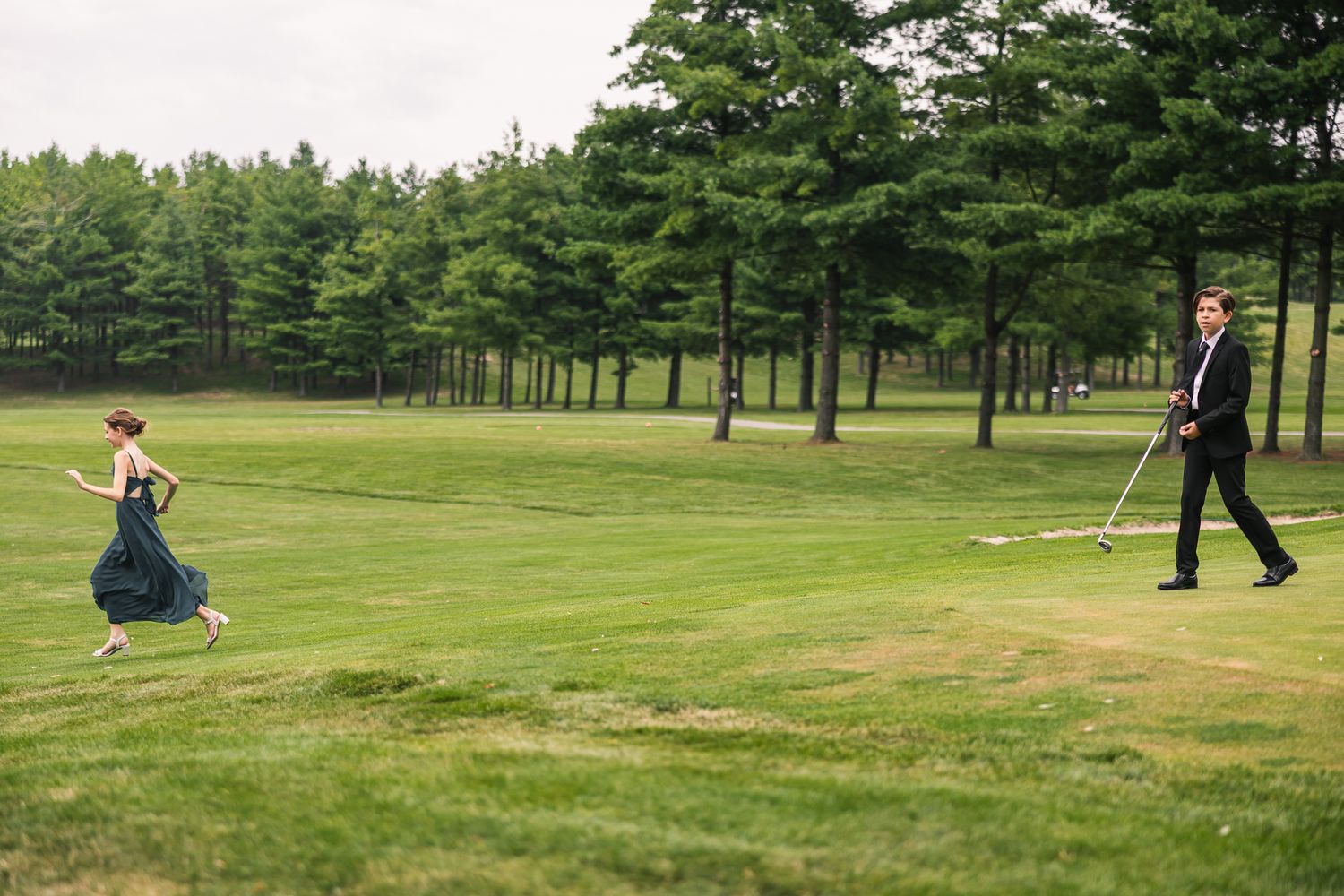 Wedding guest in navy gown runs across green golf course while groomsman walks with flag.