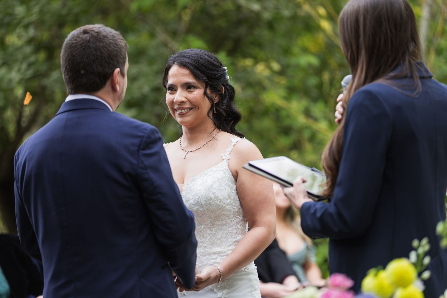 Bride smiling at groom during outdoor wedding ceremony with officiant holding book.
