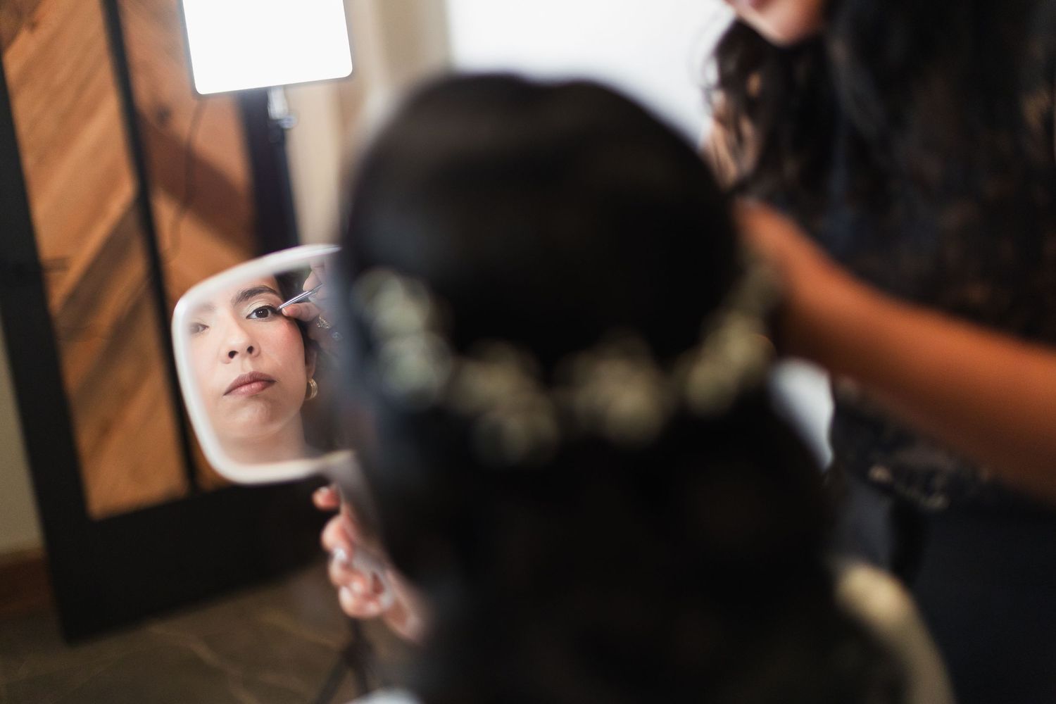 Bride getting hair styled with flower crown, reflection visible in handheld mirror during wedding preparation.