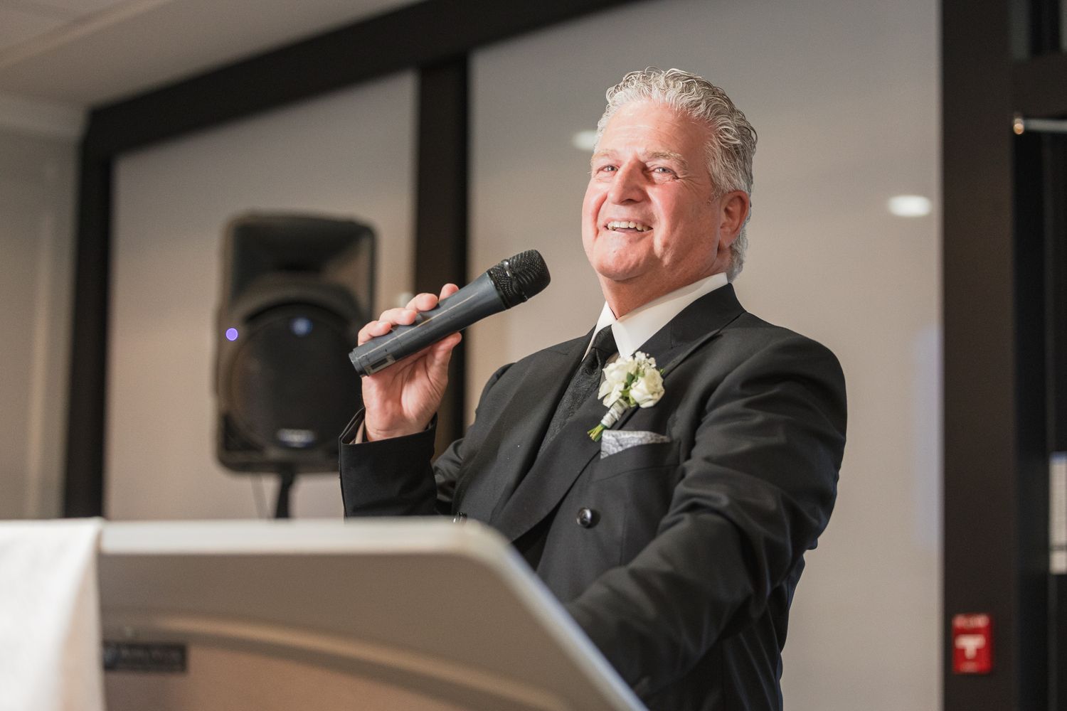 Elegant man in black tuxedo with boutonniere speaking into microphone at wedding reception podium.