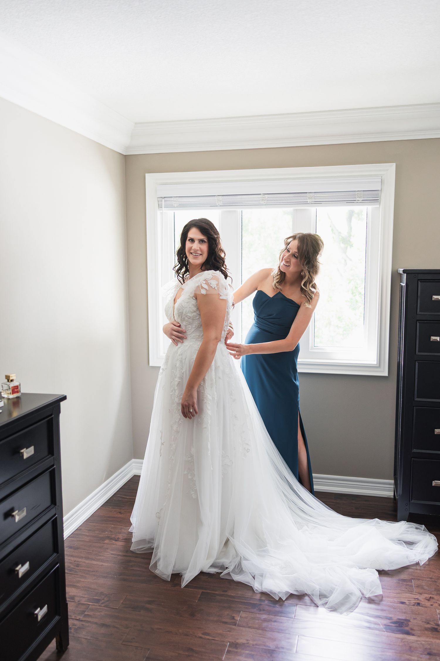 Bride in white lace gown being helped by bridesmaid in teal dress near window on wedding day.