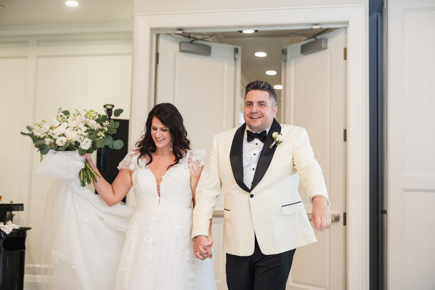 Bride and groom holding hands walking into wedding reception, bride in lace gown holding white bouquet, groom in ivory tuxedo.
