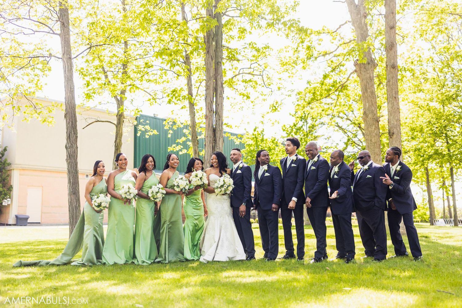 Full wedding party in navy suits and mint green dresses pose together outdoors under spring trees.