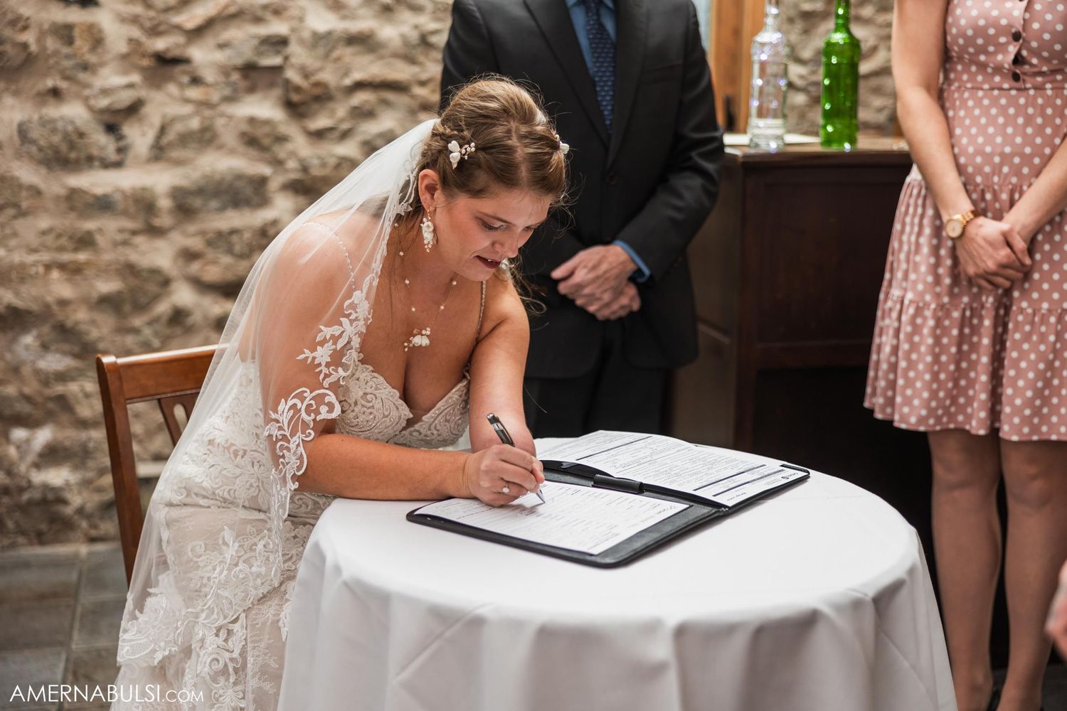 Person in wedding dress signs documents at white-clothed table in stone-walled venue.