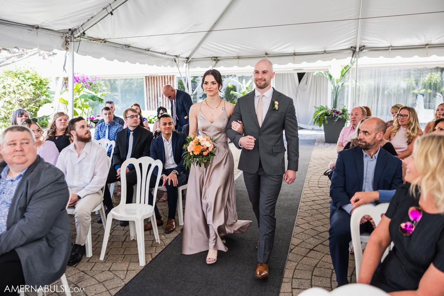 Wedding ceremony under a white tent with guests seated on white chairs.