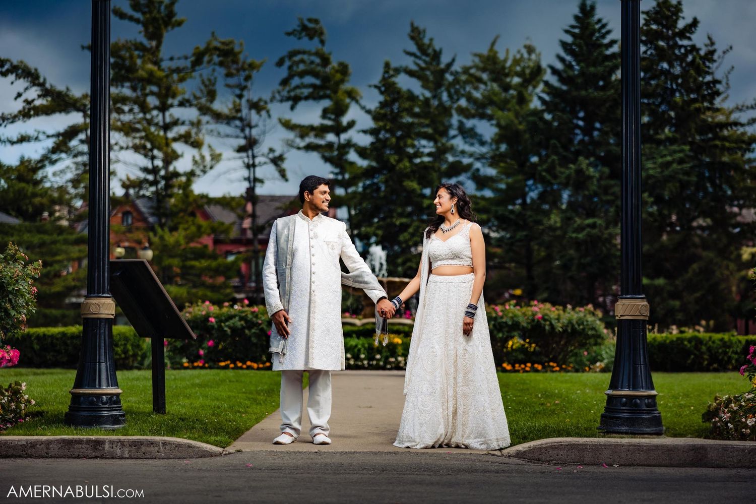 A wedding couple poses on a garden pathway with tall evergreen trees and ornate lamp posts in the background.
