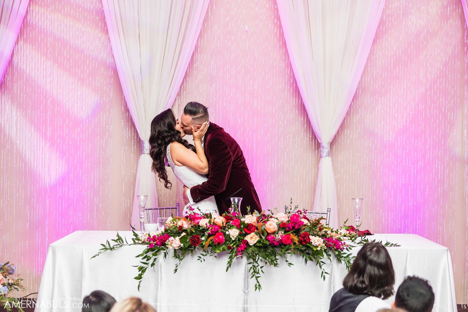 Romantic kiss at wedding reception head table decorated with red floral arrangements.