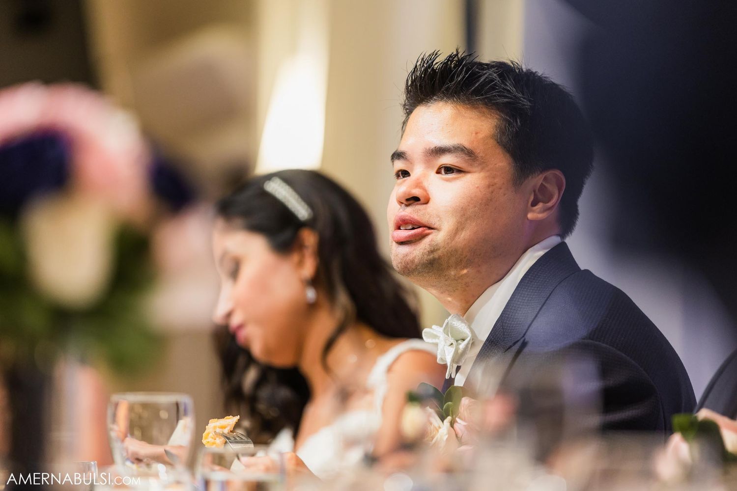 Wedding reception candid moment captured at elegant head table with floral decorations.
