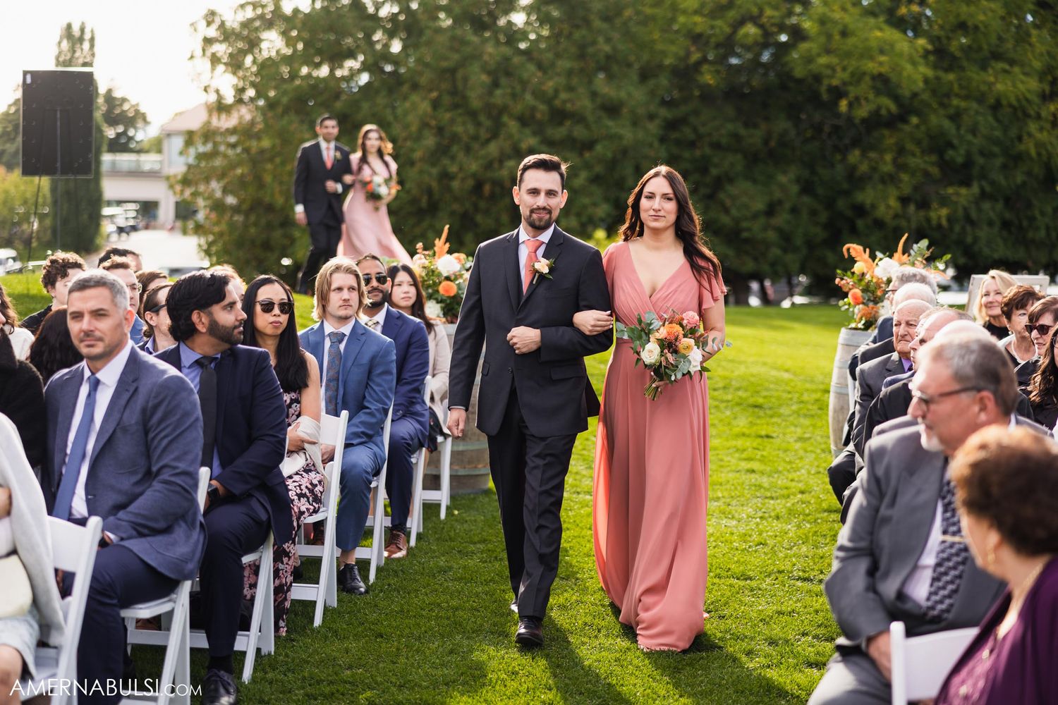 A bridesmaid in pink and groomsman process down an aisle during an outdoor wedding ceremony.